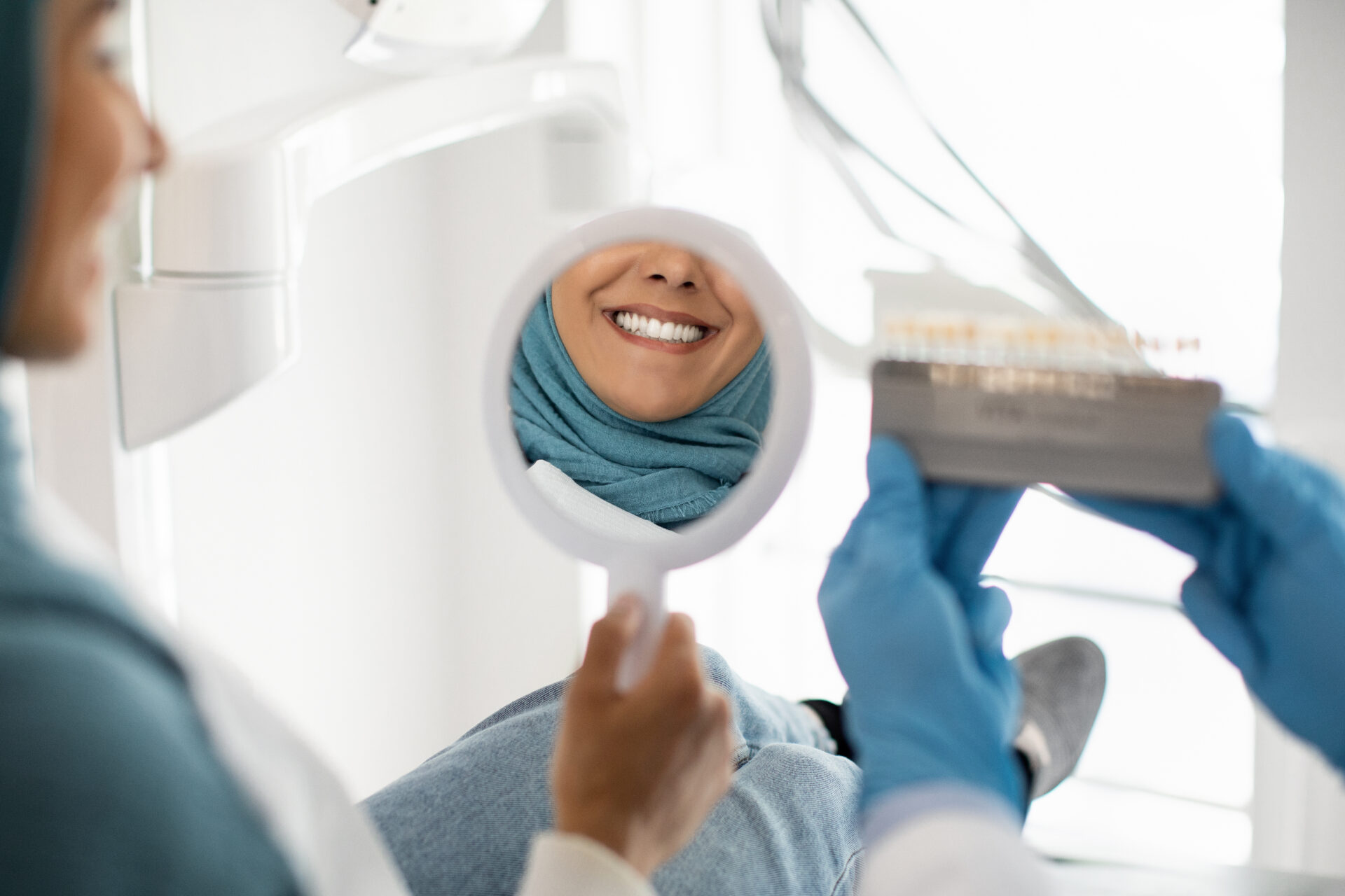 Beautiful Muslim Woman In Hijab Looking At Mirror In Dentists Office, Islamic Female Patient Enjoying Her Beautiful Smile And Teeth After Dental Treatment In Stomatologic Clinic, Selective Focus